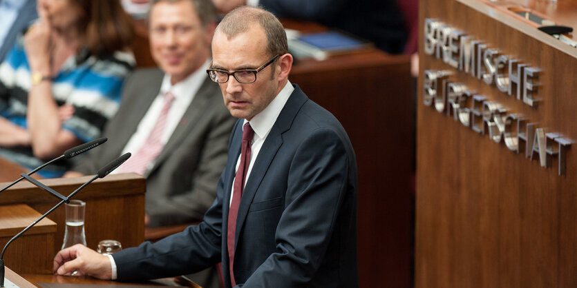 Ein mittelalter Mann mit Anzug, hoher Stirn, dunkler Brille und braunen Haaren steht hinter einem hölzernen Rednerpult.