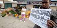 Ein Migrant steht vor einem Laden und demonstriert. Auf seinem Schild steht: "Heute arbeite ich nicht für 50 Euro am Tag".