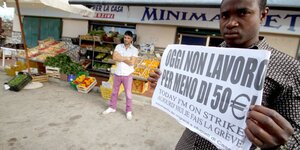 Ein Migrant steht vor einem Laden und demonstriert. Auf seinem Schild steht: 