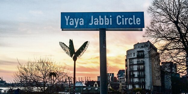 Auf einem Schild steht "Yaya Jabbi Circle" mit weißer Schrift auf blauem Grund Im Hintergrund steht eine künstliche Palme des Park Fiction.