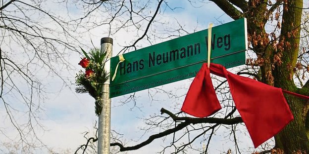 Schild "Hans-Neumann-Weg" mit rotem tuch und Blumen.