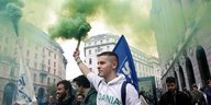 Ein junger Mann inmitten einer Menschenmenge hält eine Fackel hoch, aus der viel grüner Rauch kommt