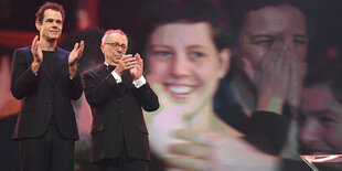 Zwei Männer applaudieren auf einer Bühne, im Hintergrund auf einer Leinwand das überlebensgroße Gesicht einer Frau