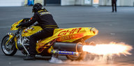 Ein Mann sitzt auf einem gelben Motorrad, aus dessen Auspuff eine Stichflamme kommt