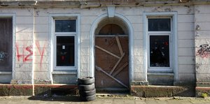 Eine Hausfassade mit aufgeplatztem Putz, Graffiti und einer mit Holzbalken vernagelten Tür.