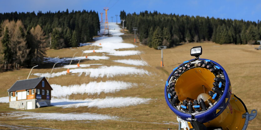 eine Skipiste die spärlich mit Kunstschnee bedeckt ist, im Vordergrund eine Schneekanone