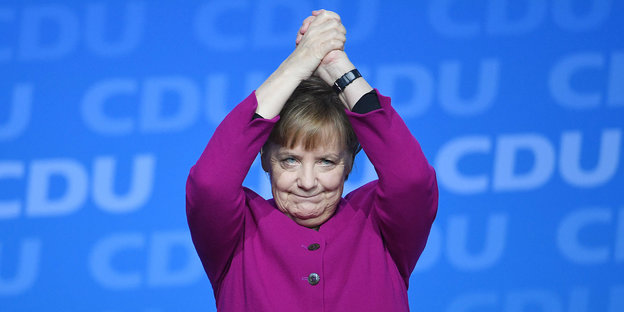 Angela Merkel steht vor einer CDU-Plakatwand und hat die Hände zu einer Siegerpose erhoben