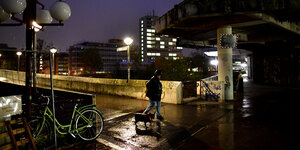 Eine Frau mit Regenjacke und Hund geht bei Dunkelheit über eine nasse, schlecht beleuchtete Betonbrücke.