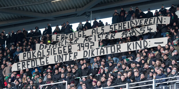 Ein mit Menschen gefüllter Zuschauerrang im Stadion von Hannover 96. Auf einem Plakat ist von "Diktator Kind" und einem "Scheißverein" die Rede.