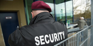 Ein kräftiger Mann lehnt an einem Geländer. Auf seiner Jacke steht "Security".