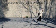 Ein alter Mann geht an einer Mauer vorbei, vor der Blumen liegen. Auf der Mauer steht "Gedenkstätte Bergen-Belsen".