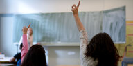 Zwei Schülerinnen strecken in der Klasse den Arm in die Höhe, einen Lehrerin steht an der Tafel