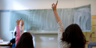 Zwei Schülerinnen strecken in der Klasse den Arm in die Höhe, einen Lehrerin steht an der Tafel