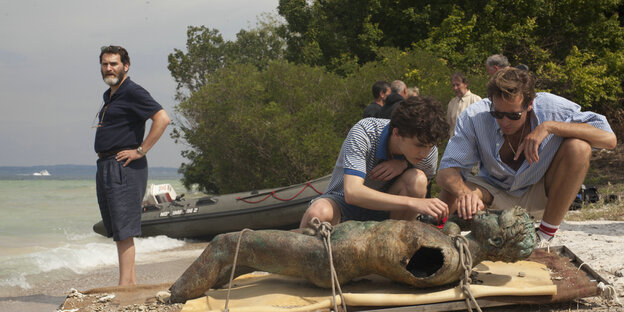 Zwei junge Männer betrachten eine männliche Statue, die auf einem Strand liegt. In der Dünung steht ein älterer Mann