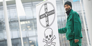 Ein Mann in grüner Kleidung und Mütze hält ein Banner, auf dem die Schriftzüge von Bayer und Monsanto sowie ein Totenschädel zu sehen sind