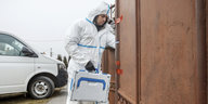 Ein Mann in weißem Schutzanzug trägt einen weißen Koffer durch eine verrostetes Tor