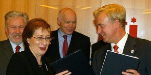 Eine Frau und ein Mann halten jeweils eine Mappe in der Hand, im Hintergrund zwei Männer in Anzügen, alle lachen.