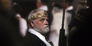 Ein Mann mit einer Krone auf dem Kopf und einem Gewehr in der Hand blickt grimmig in die Kamera