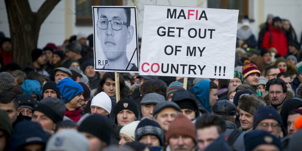 Eine Menschenmenge, aus der Schilder in die Höhe ragen. Auf einem steht „Mafia, get out of my country“