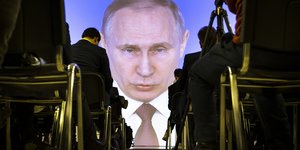 Menschen sitzen in Reihen vor einer Leinwand, auf der Putins Gesicht zu sehen ist