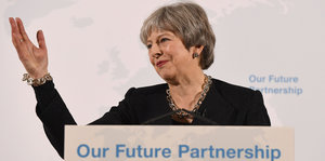 Theresa May, winkend, an einem Pult auf dem „Our Future partnership“ steht