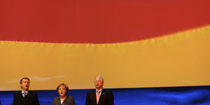 Angela Merkel singt die deutsche Nationalhymne vor einer deutschen Fahne. Neben ihr stehen zwei Parteikollegen