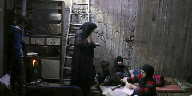 Eine Frau steht in einem Bunker und guckt zu zwei Kinder und einer Frau n, die vor ihr auf dem Boden sitzen