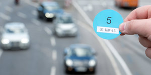 Eine Hand hält eine blaue Plakette vor eine Straße mit Autos