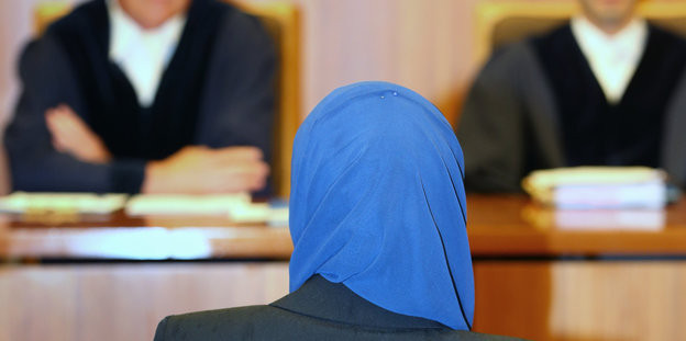 Eine Frau mit blauem Kopftuch ist von hinten zu sehen. Vor ihr sieht man unscharf einen Richter und eine weitere Person