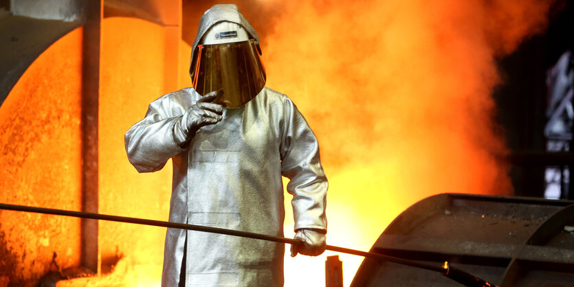 Ein Stahlarbeiter in Schutzkleidung mit Helm steht vor eine lodernden Feuer