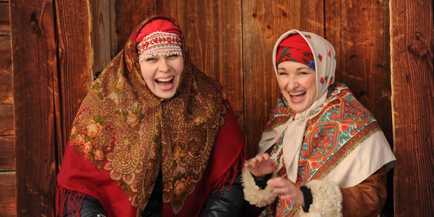 Zwei Frauen in traditionell russicher Kleidung sitzen vor einer Holzwand und lachen