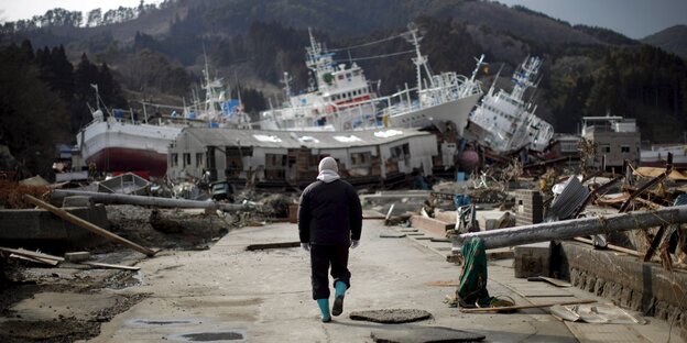 Ein Mann läuft auf einer Hafenstraße, lings und rechts zerstörte Dinge, vor ihm ein Haufen aus Schiffen und einem Haus