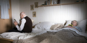 Ein alter Mann sitzt auf einem Ehebett und hält ein Telefon in der Hand. Auf der anderen Seite des Bettes liegt eine Frau mit geschlossenen Augen, die ein Blumensträuschen in den Händen hält