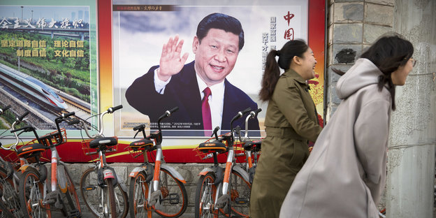 Zwei Frauen eilen an einem Plakat mit dem winkenden Staatspräsidenten Xi Jinping vorbei