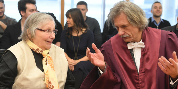 Eine ältere Dame mit Halstuch spricht mit einem Mann neben ihr, der eine rote Anwaltsrobe trägt