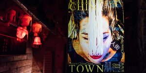 Screen mit chinesischer Frau, der Nudeln aus dem Mund hängen. Daneben rote Lampions.