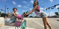 Eine junge Frau in kurzer Hose schiebt einen Einkaufswagen, in dem eine kleines Mädchen sitzt. Die Frau hat bunte Haare und streckt ihre Zunge raus