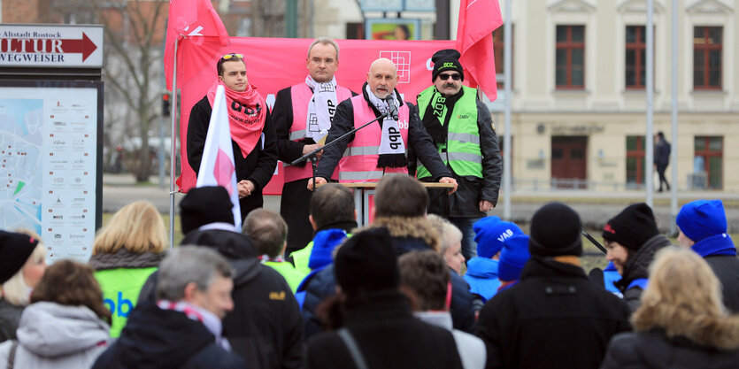 Viele Menschen haben sich an einem Platz versammelt, ein paar stehen vor roten Fahnen auf einer kleinen Bühne