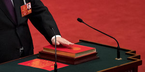 Ein rotes Buch auf einem Pult mit Mikrofon. Eine Hand liegt darauf. Man sieht nur den Arm des Mannes
