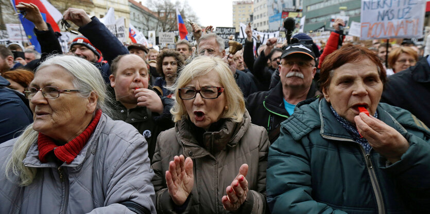 Drei Frauen stehen nebeneinander: Die mittige ruft, die an der rechten Seite bläst in eine Trillerpfeife. Hinter ihnen weitere Demonstranten