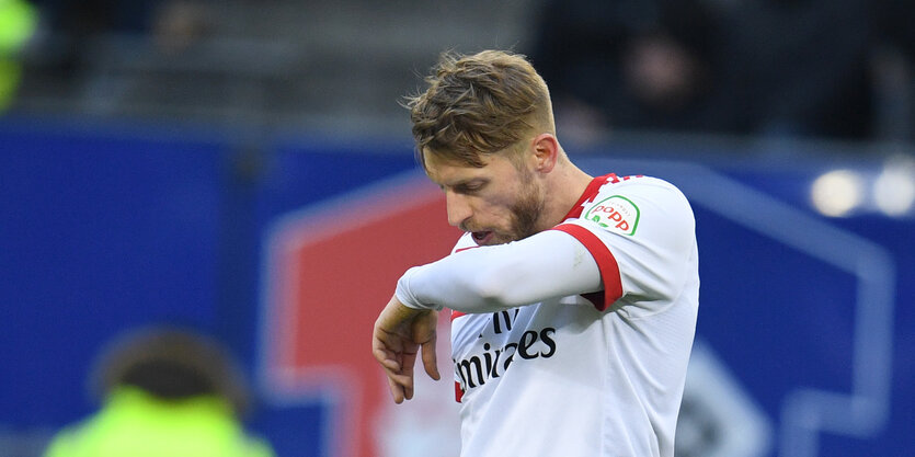 Ein junger Mann mit weiß-rotem Fußballtrikot und blonden Haaren wischt sich mit dem Arm über das Gesicht.