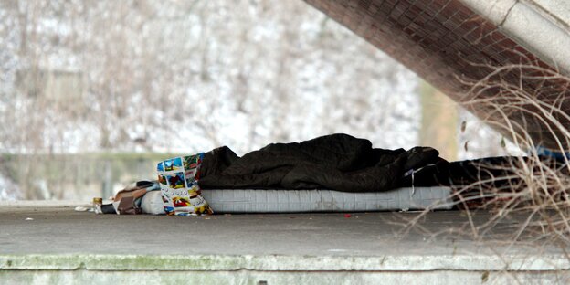 Ein Schlafsack liegt unter einer Brücke auf dem gepflasterten Boden.