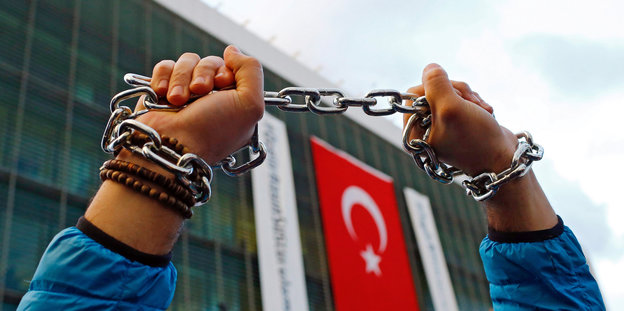 Hände, die eine Kette in die Höhe halten vor einem Gebäude, an dem die türkische Flagge hängt