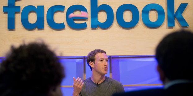 Mark Zuckerberg sitzt unter einem Facebooklogo