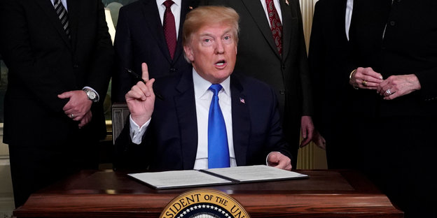 Trump sitzt an einem Schreibtisch und hält den Zeigefinger hoch