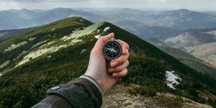 Eine Hand hält einen Kompass. Im hintergrund ist Gebirge zu sehen