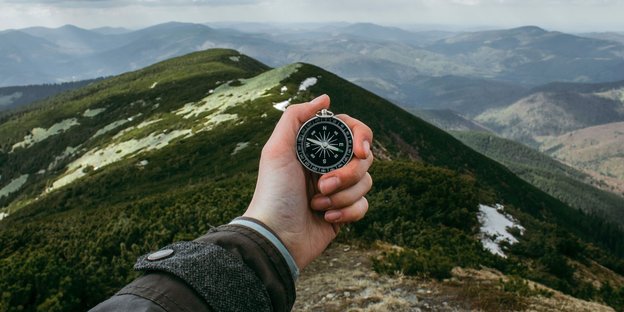 Eine Hand hält einen Kompass. Im hintergrund ist Gebirge zu sehen