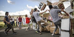 Eine Gruppe von Menschen läuft auf der Landefläche eines Flughafens gegen eine Wand aus leeren Pappkartons