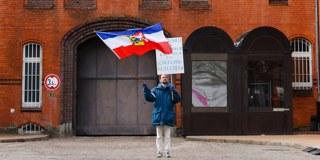 Ein Mann steht mit einer Flagge vor einem roten Backsteinhaus