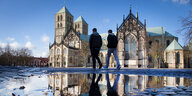 Der Dom in Münster spiegelt sich in großflächigen Pfützen, zwei Menschen schreiten über den Platz